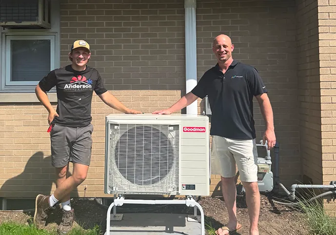 Two Anderson technicians beside a Goodman heat pump outdoor unit