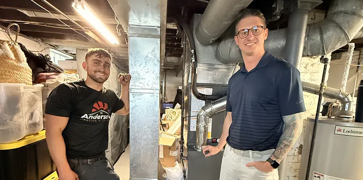 Two Anderson Heating and Cooling team members on either side of a furnace and its ductwork in a basement